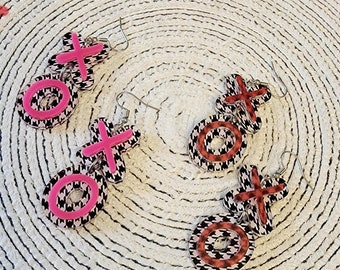 May include: Two pairs of earrings with a black and white houndstooth pattern. Each earring features a pink or red circle with a pink or red "X" or "O" on top. The earrings are on a white and black circular background.