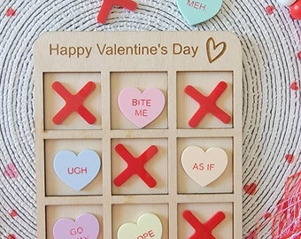 May include: A wooden tic-tac-toe game board with the words "Happy Valentine's Day" at the top. The board has nine squares, each with a red X or a heart-shaped candy with a Valentine's Day message. The candies say "Bite Me", "Ugh", "Go Away", "As If", "Nope", and "Meh".
