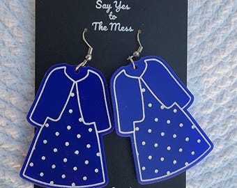 May include: A pair of blue acrylic earrings shaped like dresses with white polka dots. The earrings have a matching blue jacket design and silver-colored hooks. The earrings are displayed on a black card with white text.