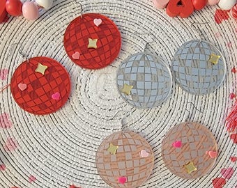 May include: A set of six disco ball earrings with different colored accents. The earrings are made of clear acrylic and have a mirrored finish. The accents are small hearts and stars in pink, red, and gold.