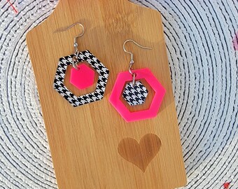 May include: A pair of pink and black geometric earrings with a houndstooth pattern. The earrings are on a wooden surface with a heart design.
