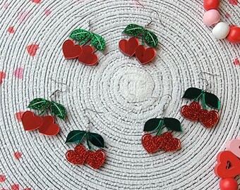 May include: Four pairs of red and green cherry earrings with glitter. The earrings are on a white background with red and pink hearts.