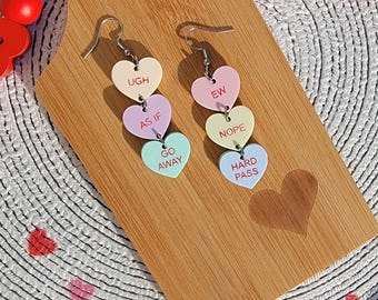 May include: A pair of earrings with three heart-shaped charms on each earring. The charms are in different colors and have the following text: "UGH", "AS IF", "GO AWAY", "EW", "NOPE", "HARD PASS".