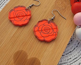 May include: A pair of red rose earrings with silver hooks. The earrings are on a wooden surface with a heart shape carved into it.