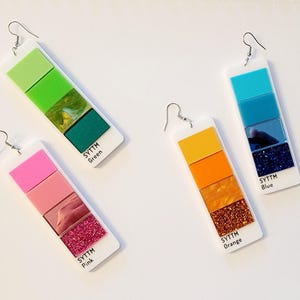 Op de afbeelding: Vier paar bungelende oorbellen met een kleurenpaletontwerp. Elke oorbel heeft een witte rechthoekige basis met een kleurengradi&euml;nt, waaronder groen, roze, oranje en blauw. De tekst "SYTTM" en een kleurnaam staan op elke basis.