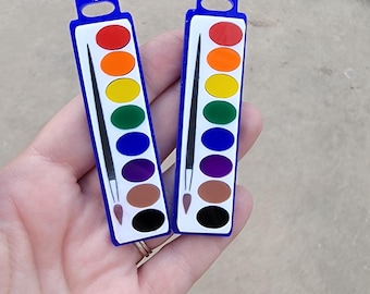 May include: Two watercolor palette-shaped earrings. Each earring has a blue border, white center, and a rainbow of colors, including red, orange, yellow, green, blue, purple, and brown. A black paintbrush is also included.