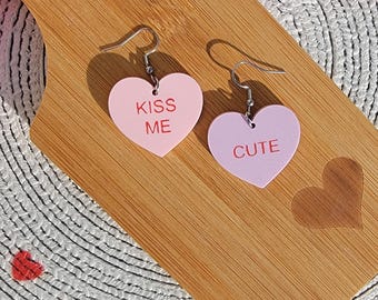 May include: A pair of pink heart-shaped earrings with the words "Kiss Me" and "Cute" on them. The earrings are on a wooden surface.