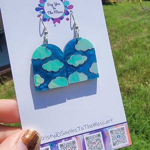May include: A pair of cloud-themed earrings with silver hooks. The earrings are arch-shaped with a blue background and white and teal cloud designs. The card behind the earrings has the text "Say Yes to The Mess" and social media QR codes.