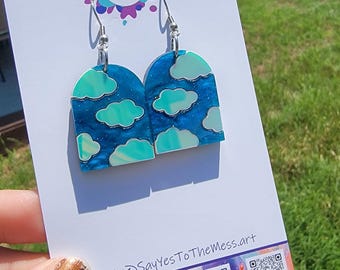 May include: A pair of cloud-themed earrings with silver hooks. The earrings are arch-shaped with a blue background and white and teal cloud designs. The card behind the earrings has the text "Say Yes to The Mess" and social media QR codes.