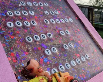 May include: A pink framed art piece with the text "MAMAS DON'T LET YOUR BABIES GROW UP TO BE ASCISTS" in white letter beads. A toy baby and yellow and orange toy truck are at the bottom. The background is filled with glitter.