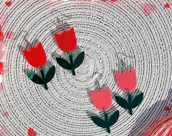 May include: A pair of red and pink acrylic tulip earrings with green leaves. The earrings are on a white background with a red and pink heart pattern.