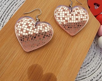 May include: A pair of rose gold heart-shaped earrings with a disco ball design. The earrings are on a wooden surface with a heart-shaped shadow.