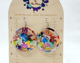 May include: A pair of colorful, round acrylic earrings with a white background and a silver hook. The earrings are displayed on a wooden stand with a ruler and the text "Say Yes to The Mess" and "Out of the mess, comes the masterpiece." The stand also includes the website address "www.SayYesToTheMess.art".