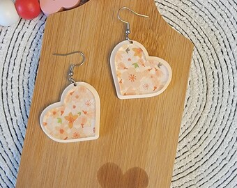 May include: A pair of heart-shaped earrings with a floral pattern on a wooden cutting board. The earrings are made of white resin with a clear coating and have silver hooks.