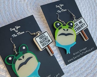 May include: Frog-shaped earrings with green and blue accents, each holding a sign. One sign reads "Frogs Against Fascism," the other "Give Me Ribberty." The earrings are on black cards with "Say Yes to The Mess" text.