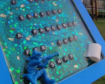May include: A blue framed art piece with the text "I DIDN'T SURVIVE A PANDEMIC FOR THIS SHIT." The artwork features a blue toy soldier aiming a rifle, set against a glittery, teal background.