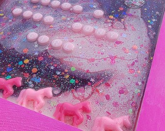 May include: A pink framed art piece with a glittery, galaxy-themed background. The artwork features pink pony figurines and the words "PINK PONY" spelled out in pink letter beads. A small cowboy hat is also included.