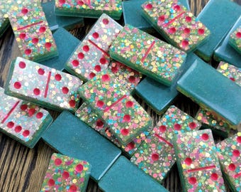 May include: A set of teal green dominoes with red dots and colorful glitter. The dominoes are scattered on a wooden surface.