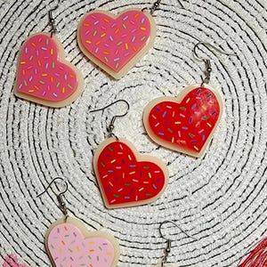 Op de afbeelding: Vier paar hartvormige oorbellen met een roze of rode glazuur en witte sprinkles. De oorbellen zijn gemaakt van polymer clay en hebben zilveren haakjes.