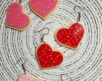 May include: Four pairs of heart-shaped earrings with a pink or red glaze and white sprinkles. The earrings are made of polymer clay and have silver hooks.