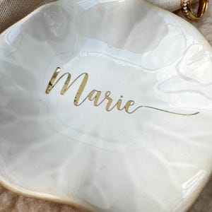 Op de afbeelding: Een witte, schelpvormige sieradenschaal met een gouden rand en de naam "Marie" in gouden letters. De schaal heeft een glanzende afwerking en een subtiel, gemarmerd patroon. Een gouden ring is zichtbaar op de achtergrond.