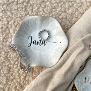 Op de afbeelding: Een witte, zeshoekige sieradenschaal met de naam "Jana" in zwart schrift. Een zilveren ring en armband liggen op de schaal. De schaal heeft een bladachtig patroon en een gouden rand. De achtergrond is een beige, getextureerd oppervlak.