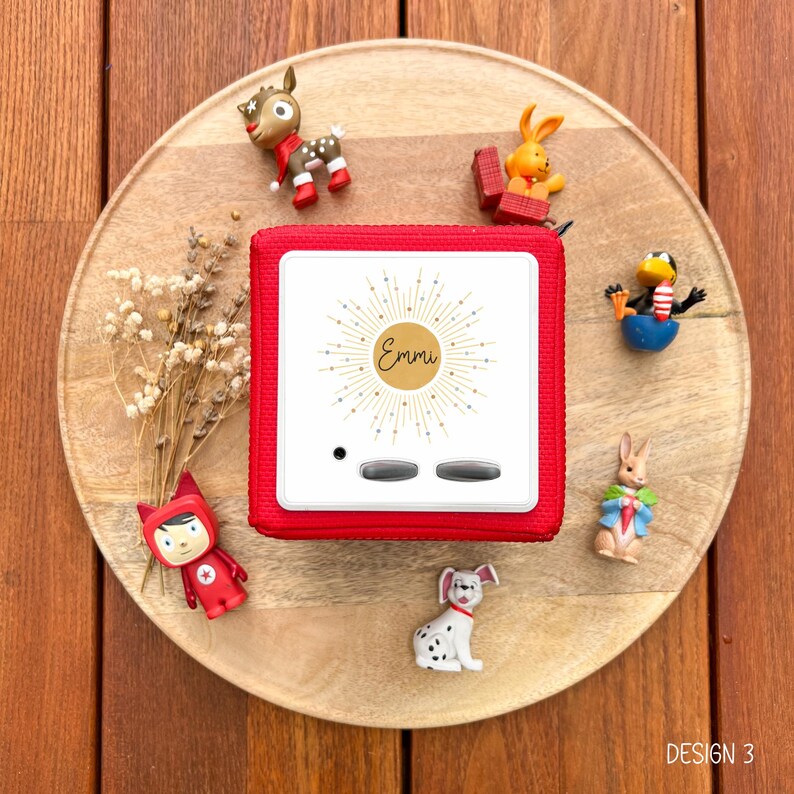 May include: A red square audio player with the name "Emmi" on the front, surrounded by a sunburst design. The player is on a wooden tray, surrounded by small figurines of animals and a character. The text "DESIGN 3" is in the bottom right corner.