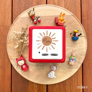 May include: A red square audio player with the name "Max" in a sun design, surrounded by small figurines of animals and a character. The player has two silver buttons and a small black dot. The items are arranged on a wooden tray.