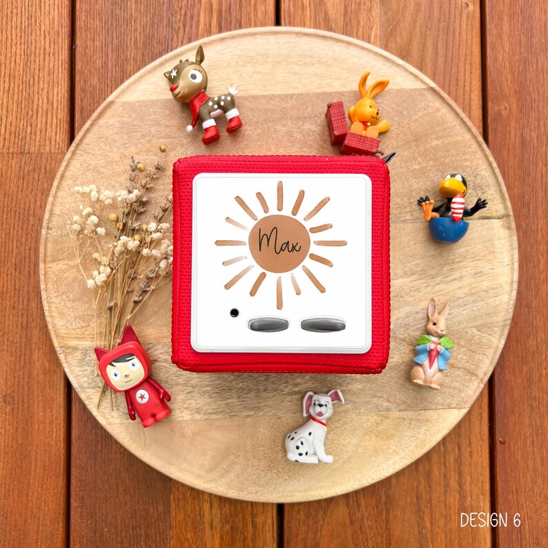 May include: A red square audio player with the name "Max" in a sun design, surrounded by small figurines of animals and a character. The player has two silver buttons and a small black dot. The items are arranged on a wooden tray.
