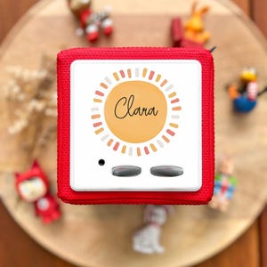 May include: A red, square-shaped audio player with a white face featuring the name "Clara" in cursive within a sunburst design. The player has two gray buttons and a small black dot. Small toy figurines surround the player on a wooden surface.