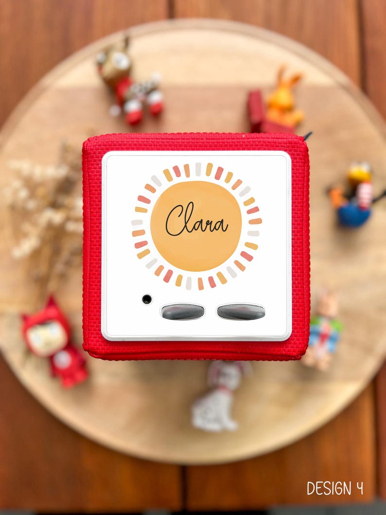 May include: A red, square-shaped audio player with a white face featuring the name "Clara" in cursive within a sunburst design. The player has two gray buttons and a small black dot. Small toy figurines surround the player on a wooden surface.