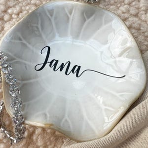 Op de afbeelding: Een witte, onregelmatig gevormde keramische schaal met de naam "Jana" in zwart cursief schrift. De schaal heeft een getextureerd, gemarmerd uiterlijk met een lichtbeige rand. Een zilverkleurige armband is gedeeltelijk zichtbaar aan de linkerkant.