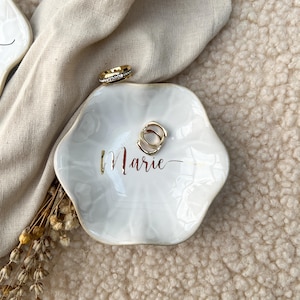 Op de afbeelding: Een witte, zeshoekige sieradenschaal met gouden rand en de naam "Marie" in gouden letters. Twee gouden ringen en een gouden en diamanten ring liggen op de schaal. De schaal ligt op een beige ondergrond.