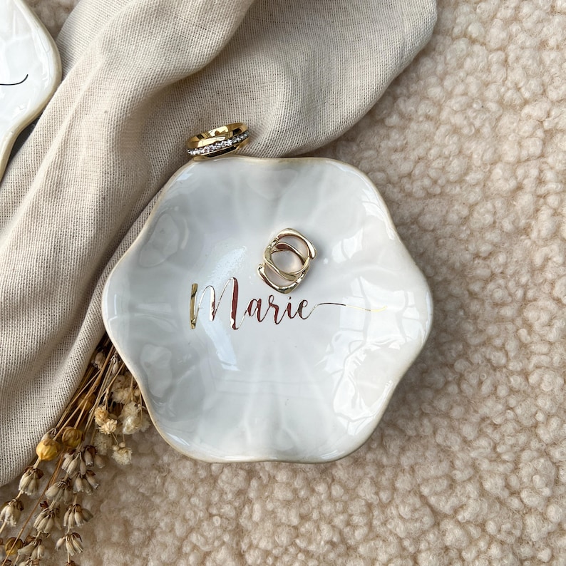 Op de afbeelding: Een witte, zeshoekige sieradenschaal met gouden rand en de naam "Marie" in gouden letters. Twee gouden ringen en een gouden en diamanten ring liggen op de schaal. De schaal ligt op een beige ondergrond.