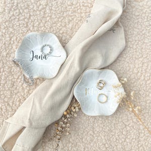 Op de afbeelding: Twee witte, bloemvormige ringhouders met gepersonaliseerde namen in script. E&eacute;n schaal heeft de naam "Jana" en een zilveren ring, de andere heeft de naam "Mia" en gouden ringen. Een beige doek en droogbloemen zijn ook aanwezig.