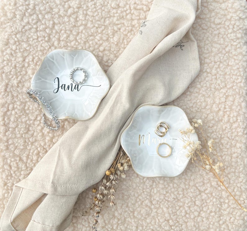 Op de afbeelding: Twee witte, bloemvormige ringhouders met gepersonaliseerde namen in script. E&eacute;n schaal heeft de naam "Jana" en een zilveren ring, de andere heeft de naam "Mia" en gouden ringen. Een beige doek en droogbloemen zijn ook aanwezig.