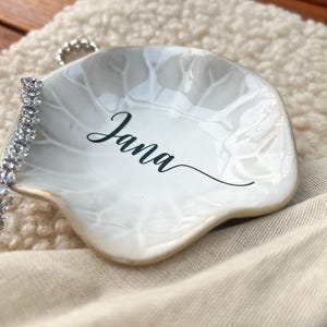Op de afbeelding: Een witte, bladvormige sieradenschaal met de naam "Jana" in zwart schrift. Een zilveren armband met heldere stenen rust op de schaal. De schaal heeft een parelmoer afwerking en een gouden rand.