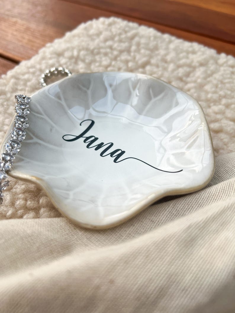 Op de afbeelding: Een witte, bladvormige sieradenschaal met de naam "Jana" in zwart schrift. Een zilveren armband met heldere stenen rust op de schaal. De schaal heeft een parelmoer afwerking en een gouden rand.