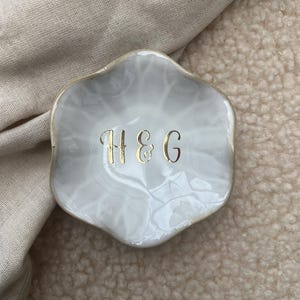 Op de afbeelding: Een kleine, wit en grijze decoratieve schaal met een gouden rand. De schaal heeft een marmer effect en heeft de gouden letters "H&G" in het midden. De schaal ligt op een beige oppervlak.