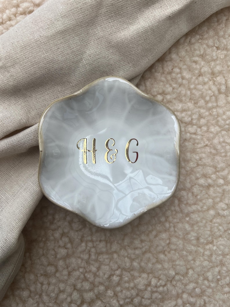 Op de afbeelding: Een kleine, wit en grijze decoratieve schaal met een gouden rand. De schaal heeft een marmer effect en heeft de gouden letters "H&G" in het midden. De schaal ligt op een beige oppervlak.