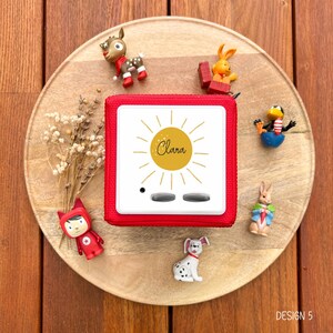 May include: A red audio player with the name "Clara" in a sun design. The player is surrounded by small figurines of animals, including a deer, rabbit, and penguin, on a wooden tray. The text "DESIGN 5" is in the bottom right corner.