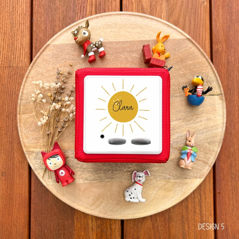 May include: A red audio player with the name "Clara" in a sun design. The player is surrounded by small figurines of animals, including a deer, rabbit, and penguin, on a wooden tray. The text "DESIGN 5" is in the bottom right corner.