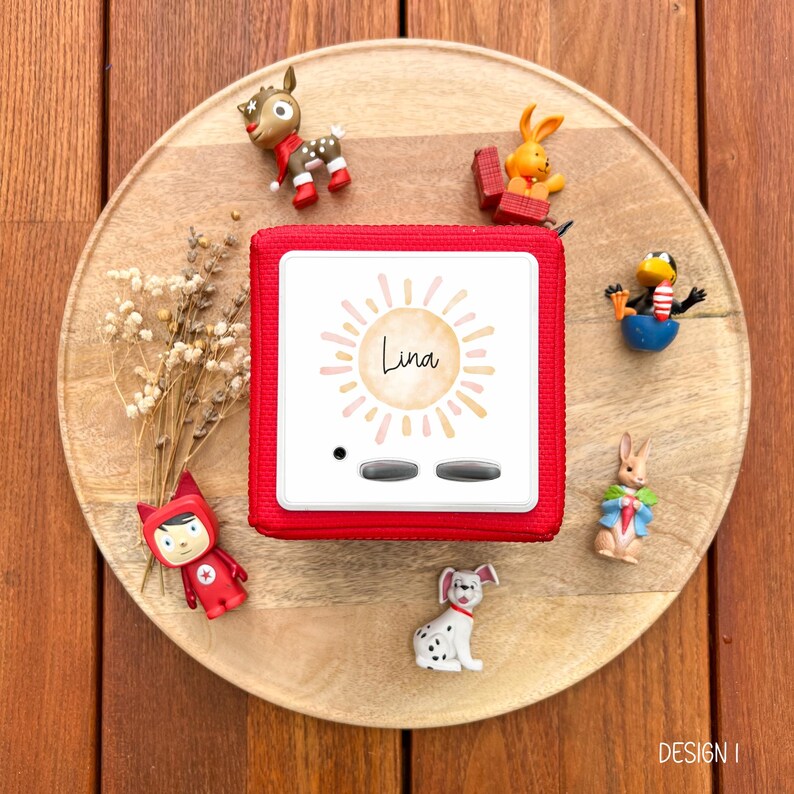 May include: A red audio player with the name "Lina" on the front, surrounded by a sun design. The player is on a wooden tray, with small figurines of animals and a red figure scattered around it. The tray is on a wooden surface.