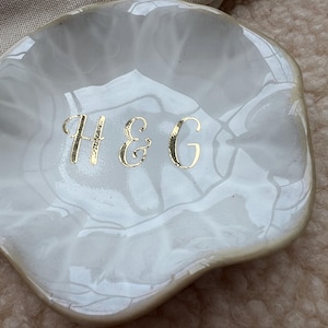 Op de afbeelding: Een witte, geschulpte keramische sieradenschaal met een gemarmerd effect. De schaal heeft gouden letters met de tekst "H & G". De schaal wordt waarschijnlijk gebruikt voor het bewaren van sieraden of kleine items.