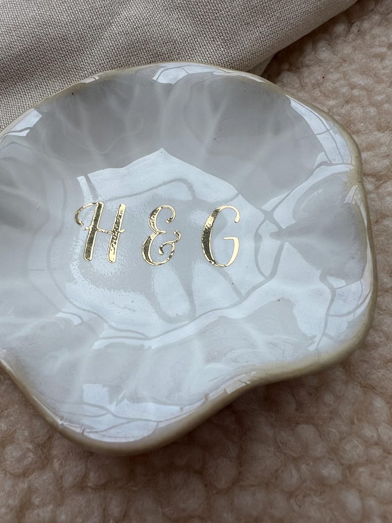 Op de afbeelding: Een witte, geschulpte keramische sieradenschaal met een gemarmerd effect. De schaal heeft gouden letters met de tekst "H & G". De schaal wordt waarschijnlijk gebruikt voor het bewaren van sieraden of kleine items.