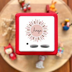 May include: A red, cube-shaped device with a white face featuring the name "Amaja" surrounded by a sunburst design. Two gray buttons and a small black dot are visible. The words "DESIGN 2" are at the bottom right.