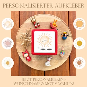 May include: A red square audio player with the name "Lina" on the front, surrounded by a sun design. The player is on a wooden tray with small figurines and decorative sun illustrations. The text at the top says "PERSONALIZED STICKER".