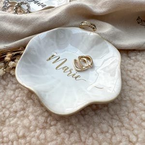 Op de afbeelding: Een witte, onregelmatig gevormde keramische sieradenschaal met gouden letters die "Marie" luidt. De schaal bevat twee gouden oorringen en een gouden ring. De achtergrond omvat een beige doek en een zachtroze oppervlak.