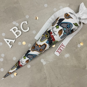 Può includere: Un cono scolastico bianco e grigio a tema videogiochi. Il cono è decorato con controller di gioco colorati, stelle e il testo "the best player". Il cono ha un nastro bianco con il nome "Alex" scritto in rosso.