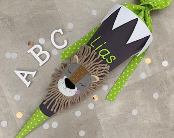 Może przedstawiać: Szary i zielony stożek szkolny z twarzą lwa i wyhaftowanym imieniem "Lias". Stożek jest ozdobiony białymi gwiazdkami i zieloną wstążką z białymi gwiazdkami.